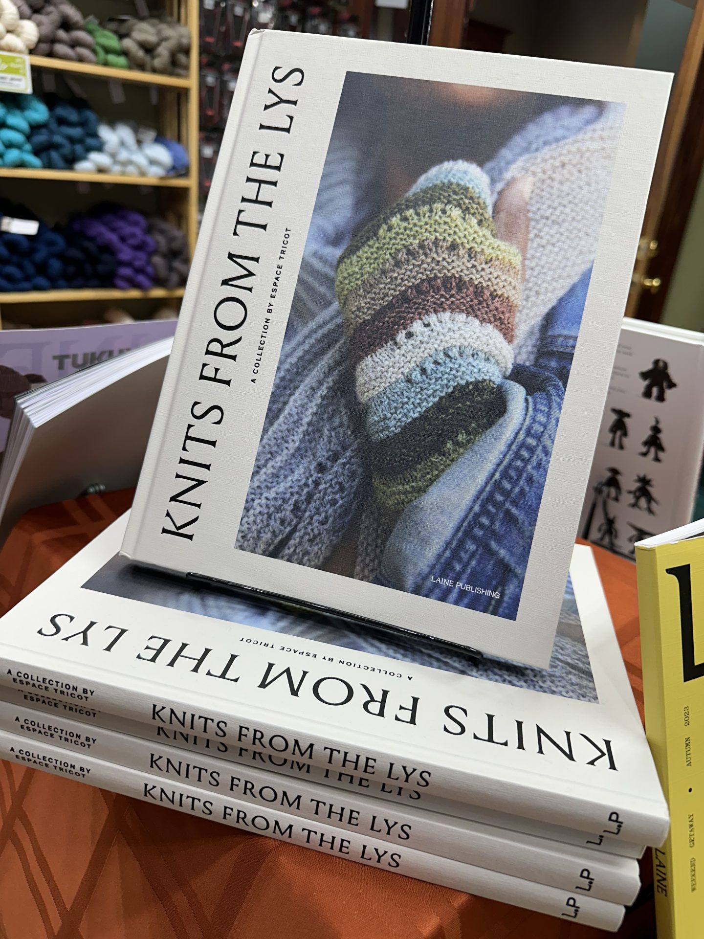 A stack of books with the title Knits from the LYS are displayed in the foreground with shelves full of yarn in the background.